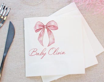May include: White paper napkins with a watercolor-style pink bow and the text "Baby Olivia". The napkins are stacked on a light-colored surface, with a fork and knife visible in the background. Ideal for a baby shower or other special event.