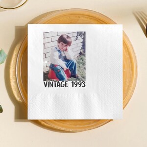 May include: White square paper napkin with a printed photograph of a young child in denim overalls, sitting on a red object. The text "VINTAGE 1993" is printed in black below the photo. The napkin is on a clear plate.