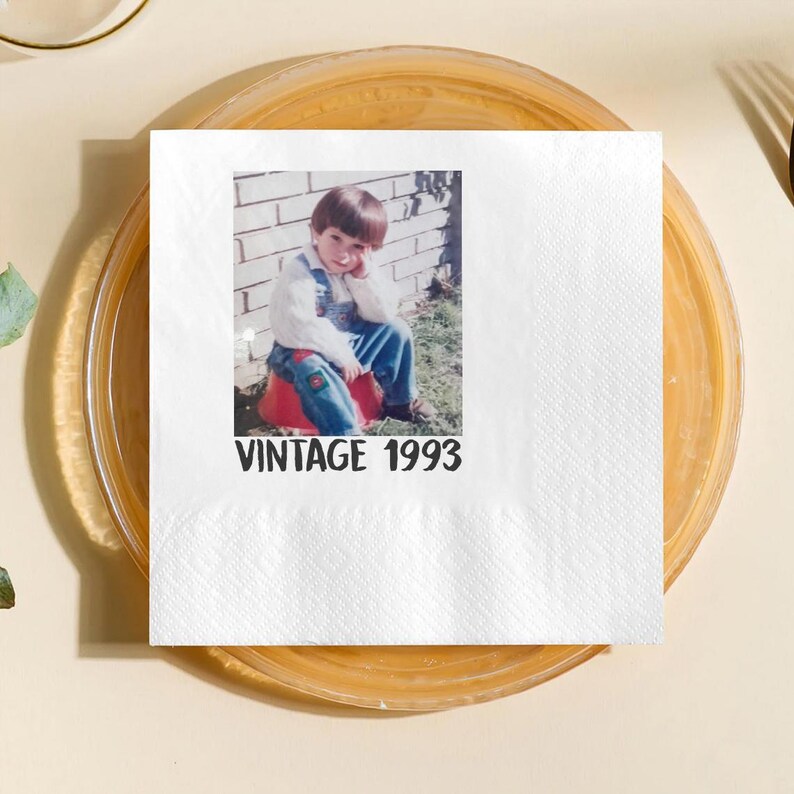May include: White square paper napkin with a printed photograph of a young child in denim overalls, sitting on a red object. The text "VINTAGE 1993" is printed in black below the photo. The napkin is on a clear plate.