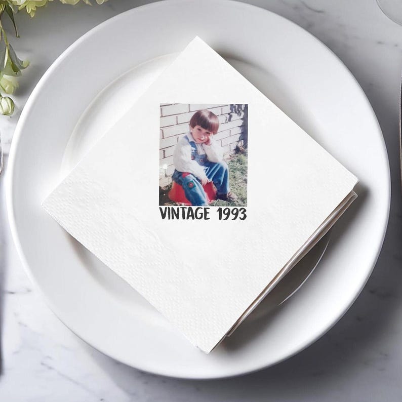 May include: White square napkin with a printed photograph of a young child in denim overalls, sitting on a red object. The text "VINTAGE 1993" is printed below the photo. The napkin is placed on a white plate.