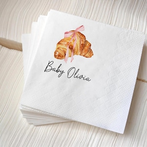 K&ouml;nnte beinhalten: Wei&szlig;e Papierservietten mit einer Aquarellillustration eines Croissants, das mit einem rosa Band gebunden ist. Der Text "Baby Olivia" ist in schwarzer Schrift unter dem Croissant-Bild geschrieben. Die Servietten sind gestapelt.