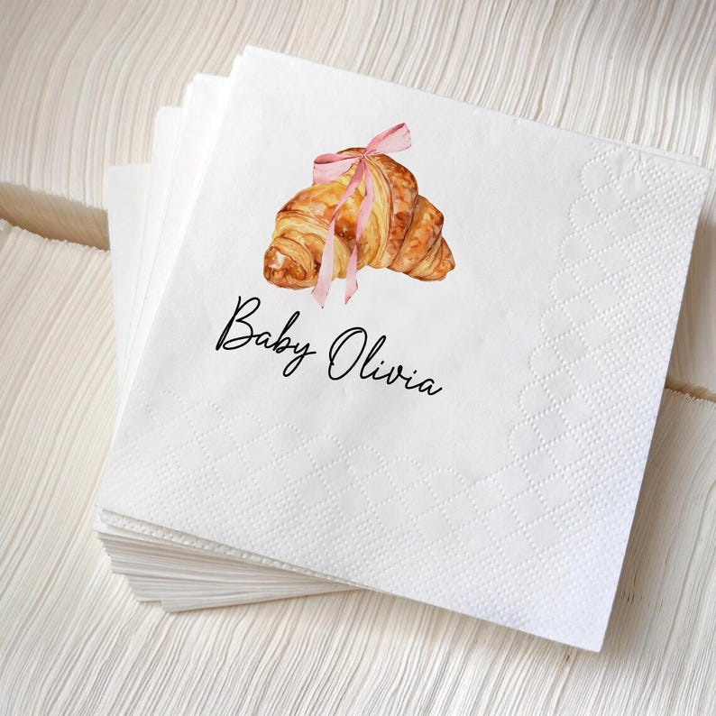 K&ouml;nnte beinhalten: Wei&szlig;e Papierservietten mit einer Aquarellillustration eines Croissants, das mit einem rosa Band gebunden ist. Der Text "Baby Olivia" ist in schwarzer Schrift unter dem Croissant-Bild geschrieben. Die Servietten sind gestapelt.