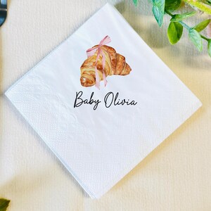 K&ouml;nnte beinhalten: Quadratische wei&szlig;e Papierserviette mit einer Aquarellillustration eines Croissants, das mit einem rosa Band gebunden ist. Der Text "Baby Olivia" ist in schwarzer Schrift unter dem Croissant geschrieben. Die Serviette hat eine strukturierte Oberfl&auml;che.