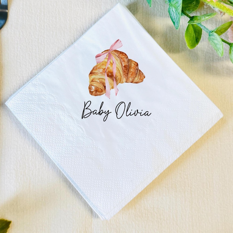 K&ouml;nnte beinhalten: Quadratische wei&szlig;e Papierserviette mit einer Aquarellillustration eines Croissants, das mit einem rosa Band gebunden ist. Der Text "Baby Olivia" ist in schwarzer Schrift unter dem Croissant geschrieben. Die Serviette hat eine strukturierte Oberfl&auml;che.