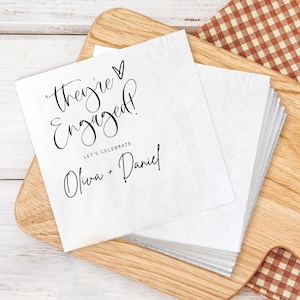 May include: White paper napkins with the text "They're Engaged!" in elegant script, along with "Let's Celebrate" and the names "Oliva + Daniel." The napkins are stacked on a wooden cutting board.