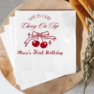 May include: White paper napkins with a red cherry design and text that reads "She is our Cherry On Top" and "Olivia's First Birthday". The napkins are stacked on a wooden tray.