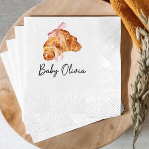 K&ouml;nnte beinhalten: Wei&szlig;e Papierservietten mit einer Aquarellillustration eines Croissants, das mit einem rosa Band gebunden ist. Der Text "Baby Olivia" ist in schwarzer Schrift unter dem Croissant geschrieben. Die Servietten sind auf einem Holztablett gestapelt.