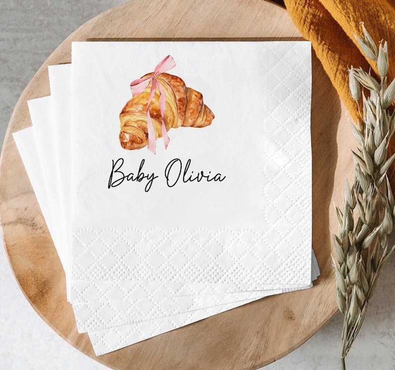 K&ouml;nnte beinhalten: Wei&szlig;e Papierservietten mit einer Aquarellillustration eines Croissants, das mit einem rosa Band gebunden ist. Der Text "Baby Olivia" ist in schwarzer Schrift unter dem Croissant geschrieben. Die Servietten sind auf einem Holztablett gestapelt.
