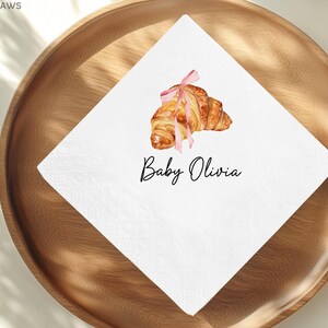 K&ouml;nnte beinhalten: Wei&szlig;e Papierserviette mit einer Aquarellillustration eines Croissants, das mit einem rosa Band gebunden ist. Der Text "Baby Olivia" ist in schwarzer Schrift darunter geschrieben. Die Serviette liegt auf einem Holztablett.