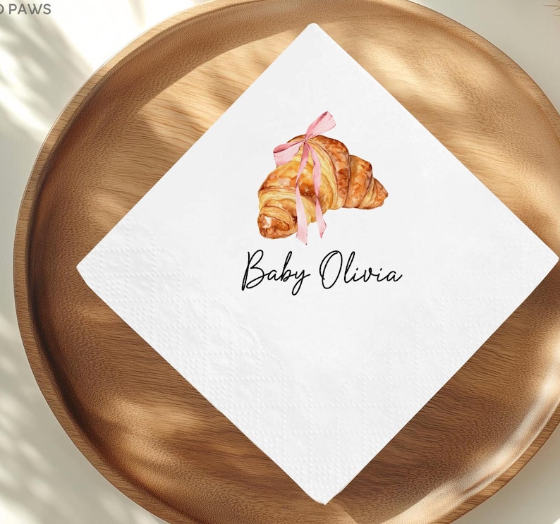 K&ouml;nnte beinhalten: Wei&szlig;e Papierserviette mit einer Aquarellillustration eines Croissants, das mit einem rosa Band gebunden ist. Der Text "Baby Olivia" ist in schwarzer Schrift darunter geschrieben. Die Serviette liegt auf einem Holztablett.