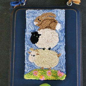 May include: A blue wooden frame with a light blue background and a needlepoint design of a brown rabbit, a black and white sheep, and a white sheep. The needlepoint is surrounded by a green border with pink and yellow flowers. A green knob is attached to the bottom of the frame.