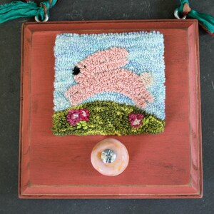 May include: A red wooden frame with a pink bunny rabbit jumping over a green hill with pink flowers. The frame has a knob on the bottom and a teal blue ribbon attached to the top.