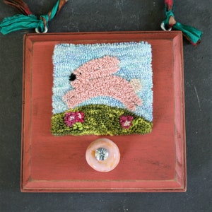 May include: A red wooden frame with a pink bunny rabbit jumping over a green hill with pink flowers. The frame has a knob on the bottom and a teal blue ribbon attached to the top.