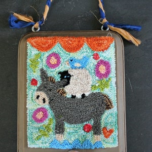 Puede incluir: Un marco de madera marrón con un diseño de punto de aguja colorido de un burro que lleva una oveja y un pájaro azul. El marco tiene un pomo marrón en la parte inferior.