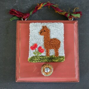 May include: A framed needlepoint artwork featuring a brown alpaca standing in a field of green grass with two red flowers. The frame is painted red and has a brown ceramic knob.