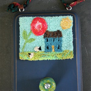 May include: A blue wooden frame with a hand-stitched needlepoint design of a house, flower, and sun. The frame has a green knob.