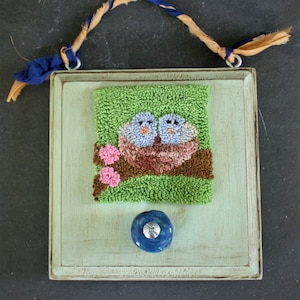 May include: A green and white framed wall hanging with a needlepoint design of two blue birds in a nest. The frame has a blue knob in the center.