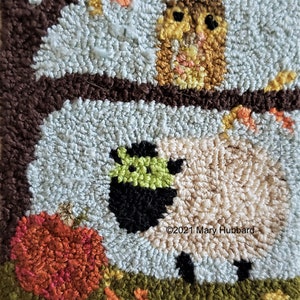 May include: A close-up of a rug hooking with a black sheep with a green face, a pumpkin, and an owl in a tree. The rug is made with different colored yarn and is a light blue background.