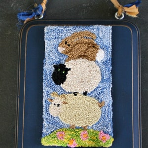 May include: A blue wooden frame with a white and brown needlepoint design of a bunny, a sheep, and a lamb. The needlepoint is framed with a blue border and has a green knob on the bottom.
