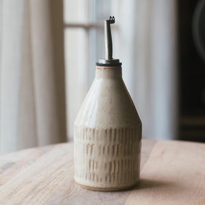 May include: A beige ceramic oil bottle with a stainless steel spout. The bottle has a textured, vertical-line design on the lower portion. It sits on a wooden table, with a blurred background of a window and a globe.