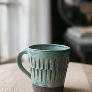 May include: A handcrafted ceramic mug with a teal glaze and a dark brown base. The mug features a textured, vertical pattern and a curved handle. The mug is sitting on a light-colored wooden surface.