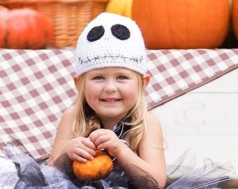 Op de afbeelding: Een jong meisje draagt een witte gehaakte muts met een Jack Skellington gezicht, een zwarte tule rok en zwart-witte schedel-gepatroneerde kniekousen. Ze zit op een baal hooi met een klein gehaakt spook speelgoed en een pompoen op haar schoot. De achtergrond is een laadbak van een vrachtwagen gevuld met pompoenen en hooi.