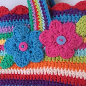 Puede incluir: Una bolsa de crochet colorida con un dise&ntilde;o de rayas arco&iacute;ris y dos flores de crochet con botones en el centro. La bolsa tiene un borde superior festoneado.
