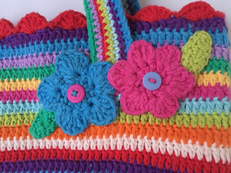 Puede incluir: Una bolsa de crochet colorida con un dise&ntilde;o de rayas arco&iacute;ris y dos flores de crochet con botones en el centro. La bolsa tiene un borde superior festoneado.
