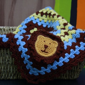 May include: A brown, blue, and yellow crocheted baby blanket with a crocheted lion face in the center. The blanket is in a wicker basket.