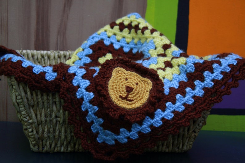 May include: A brown, blue, and yellow crocheted baby blanket with a crocheted lion face in the center. The blanket is in a wicker basket.