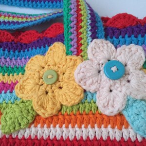 Puede incluir: Una bolsa de crochet colorida con un dise&ntilde;o de rayas arco&iacute;ris y dos flores de crochet con botones en el centro. La bolsa tiene un borde festoneado.