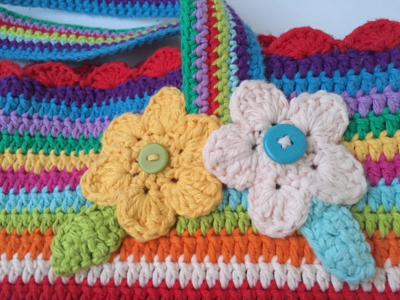 Puede incluir: Una bolsa de crochet colorida con un dise&ntilde;o de rayas arco&iacute;ris y dos flores de crochet con botones en el centro. La bolsa tiene un borde festoneado.