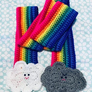 May include: Two crocheted rainbow scarves with a white crocheted cloud with a happy face and a gray crocheted cloud with a frowning face.