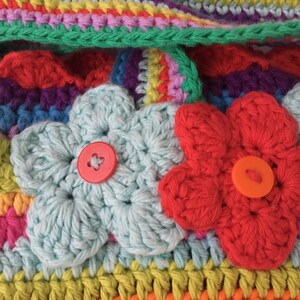 Puede incluir: Una bolsa de crochet colorida con dos flores de crochet y botones. La bolsa est&aacute; hecha con una variedad de colores, incluyendo rojo, naranja, amarillo, verde, azul, morado y rosa.
