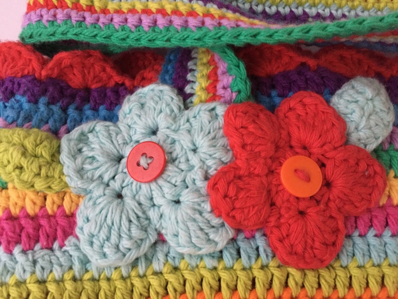 Puede incluir: Una bolsa de crochet colorida con dos flores de crochet y botones. La bolsa est&aacute; hecha con una variedad de colores, incluyendo rojo, naranja, amarillo, verde, azul, morado y rosa.