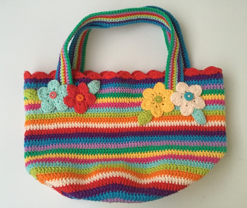 Puede incluir: Una bolsa de mano de crochet a rayas de colores con un borde festoneado y adornos florales. La bolsa tiene un arco&iacute;ris de colores, incluyendo rojo, naranja, amarillo, verde, azul, morado y rosa.