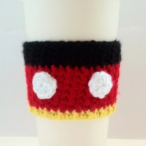 May include: A white coffee cup with a crocheted red, yellow, and black coffee cozy with white circles. The cozy has a Mickey Mouse design.