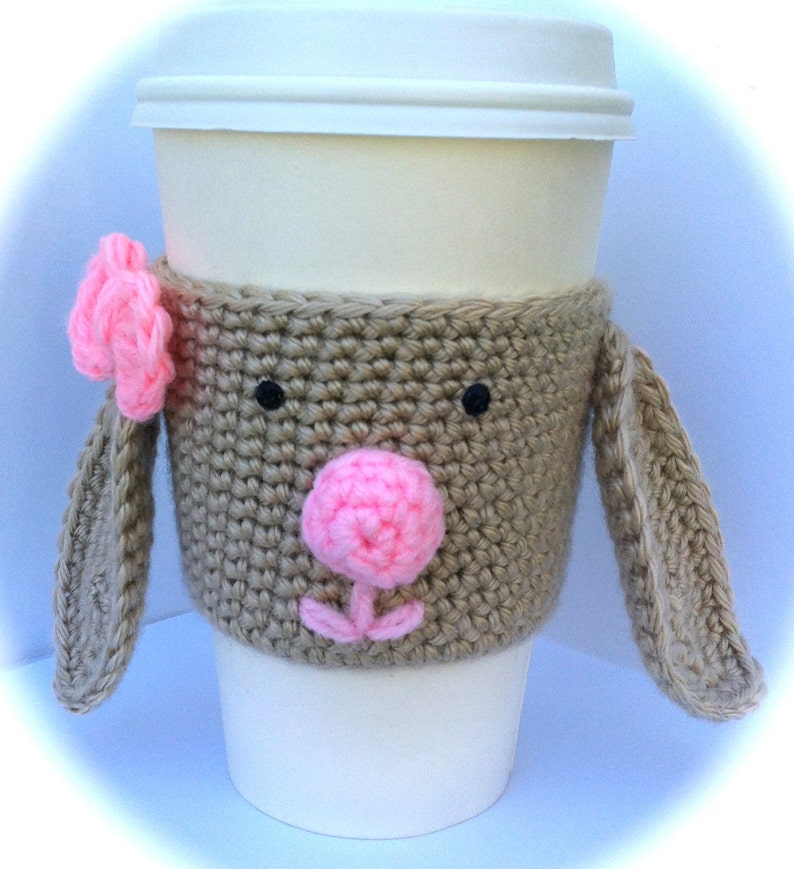 Op de afbeelding: Een gehaakte koffiebekerwarmer in de vorm van een bruin konijn met roze oren en neus. De bekerwarmer heeft zwarte ogen en een roze bloem op het oor.