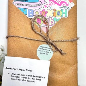 May include: A brown paper-wrapped book tied with twine, featuring colorful stickers that read I READ PAST MY BEDTIME and BOOKISH BABE. A circular sticker states it was thrifted in Wichita, KS. A white card details the psychological thriller genre.