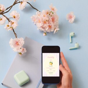 May include: A smartphone screen shows a birthday invitation with a lemon illustration and the words "Freshly 21." The phone is held in a hand, against a light blue backdrop. Pink cherry blossom branches and a notebook are also visible.