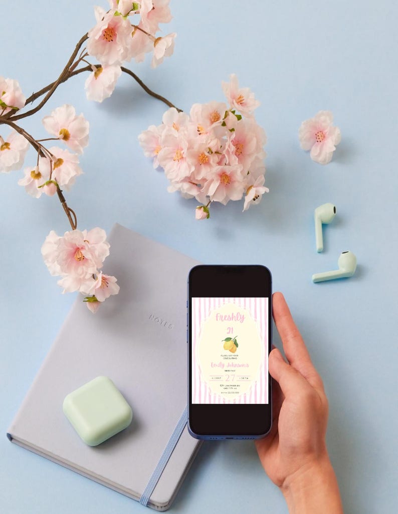 May include: A smartphone screen shows a birthday invitation with a lemon illustration and the words "Freshly 21." The phone is held in a hand, against a light blue backdrop. Pink cherry blossom branches and a notebook are also visible.