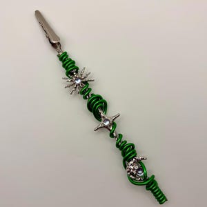 May include: A green wire clip adorned with silver-toned celestial charms. The clip features a sun, star, and crescent moon design. The wire is wrapped in a spiral pattern, creating a unique and decorative accessory.