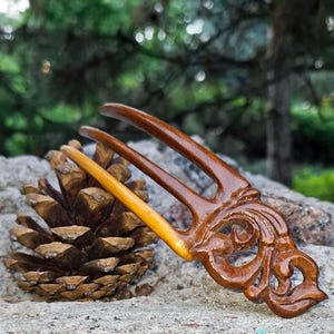 May include: A handcrafted wooden hair fork with a decorative floral design. The fork is a rich brown color with a lighter, natural wood accent. A pine cone rests beside the hair accessory, set against a natural stone background.