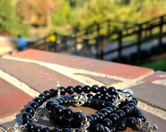 May include: Black beaded bracelets with silver-colored skull and skeleton charms are displayed on a brick surface. The bracelets are made of round, glossy black beads. The background is blurred, showing greenery and a fence.