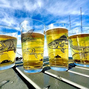 May include: Four clear pint glasses filled with a golden liquid, each featuring a different etched fish design. The glasses are arranged on a gray surface against a backdrop of a blue sky with clouds and a snowy landscape.