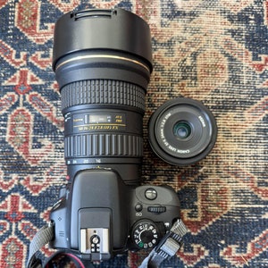 Op de afbeelding: Een zwarte Canon DSLR-camera met een zwarte lens bevestigd. De lens heeft een gouden band eromheen en de tekst "SD 16-28 F2.8 (IF) FX" erop gedrukt. Een zwarte lensdop zit op de lens. Een zwarte Canon-lens is ook in de afbeelding.