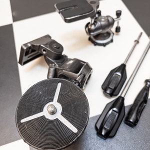 Puede incluir: Una colección de equipos de cámara antiguos, que incluyen cabezales de trípode, herramientas de ajuste y una base circular con un diseño de tres puntas. Los artículos son de metal y están dispuestos sobre una superficie a cuadros en blanco y negro.