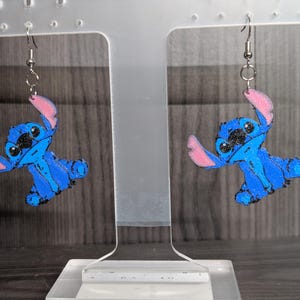 May include: A pair of blue and black Stitch earrings with pink accents. The earrings feature a cartoon character design and are suspended from silver-coloured hooks. The earrings are displayed on a clear stand against a blurred background.