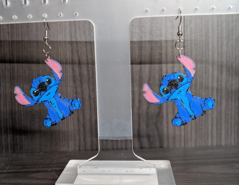 May include: A pair of blue and black Stitch earrings with pink accents. The earrings feature a cartoon character design and are suspended from silver-coloured hooks. The earrings are displayed on a clear stand against a blurred background.
