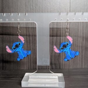 May include: A pair of blue and pink Stitch earrings. Each earring features a cartoon character design with blue body, pink ears, and black eyes. The earrings are displayed on a clear acrylic stand against a grey background.